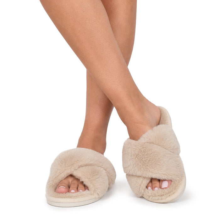 En person visar upp beige vintertofflor med mjukt foder och står med korslagda ben mot en vit bakgrund.