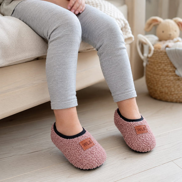 En tjej sitter på en säng i grå leggings och rosa halkfria tofflor; en korg syns i bakgrunden.
