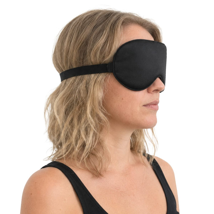 En kvinna med blont hår och svart linne bär en silkesögonmask från Kroppsbutiken och vänder sig åt höger mot en vit bakgrund.