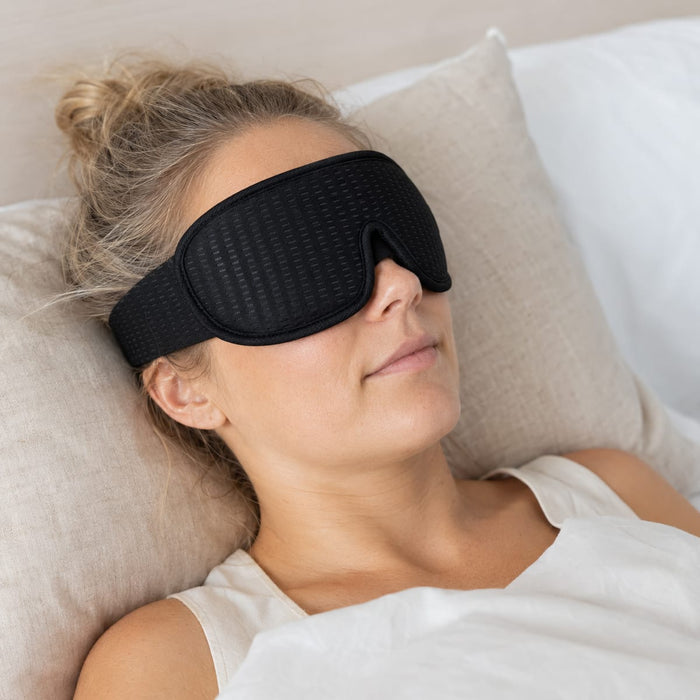 En kvinna ligger i sängen med en 3D-sömnmask från Kroppsbutiken på sig, vilar mot en beige kudde och har ett ljust täcke över sig.