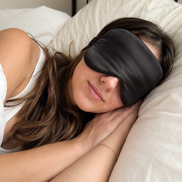 En kvinna vilar i sängen med en ögonmask i siden från Kroppsbutiken, med händerna under kinden på kudden.