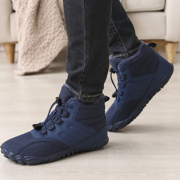 En person med fodrade vinter-Barfotaskor och mörka jeans står på ett ljust trägolv bredvid en beige soffa.