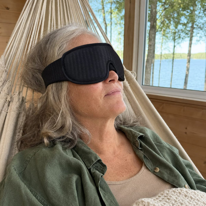 En äldre kvinna vilar i en hängmatta och bär en 3D-sömnmask från Kroppsbutiken, med utsikt över en sjö.