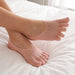 Fötter med tåstrumpor för hallux valgus vilar på en solbelyst vit säng vid ett fönster.